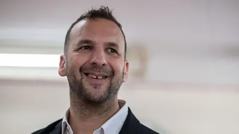 Zack Polanski looking to the side wearing a white shirt and black blazer with white backdrop