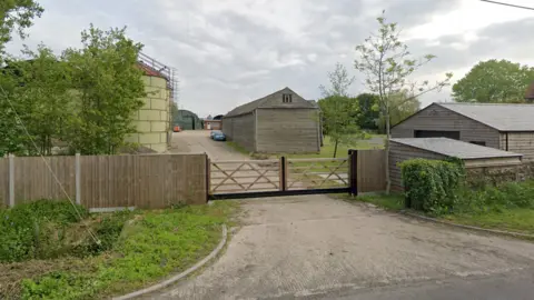 Google A fence and gate separates a road from a farm. The farm in the distance has several outbuildings and vehicles parked along a driveway. Alongside the roadside are hedges.
