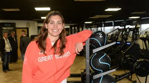 Freya Colbert stood in a gym with her arm leaning on a shelf with black weighted balls on. She has long brown hair and is smiling at the camera. She is wearing a white t-shirt and a pink zip-up jacket with white writing on it that reads "GREAT BRITAIN" in capital letters. She is also wearing grey jeans. People are stood talking behind her. 
