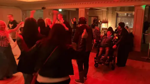 The dancefloor of the disco. It is filled with people dancing. The lights are red.