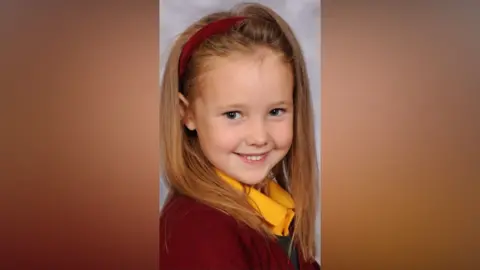 PA Elsie Dot Stancombe, has blond hair, a red alice band, yellow shirt and red school sweater.