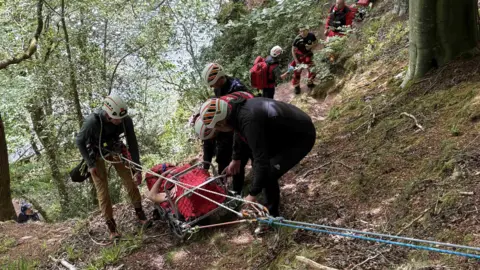 An injured man is lying on a stretcher, which is being pulled up a hill using ropes and a pulley. Four mountain rescuers in white helmets surround the man to assist him, while other rescuers stand on the path below. They are all in a wooded hilly area.