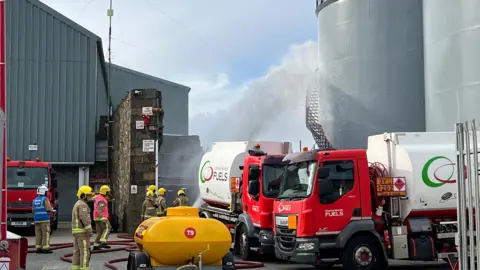 A wide shot of emergency workers gathered around fuel tankers. They are pumping water onto the tanker as if it were alight.