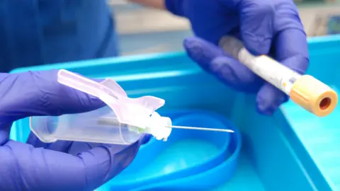 A blue gloved- hand holding a syringe with a needle, and another gloved hand holding a sample tube. They are being held over a blue tray.