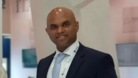 GIM Dr Vivek Koncherry smiles wearing a tie and a navy suite in an academic building. 