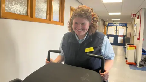 Lauren pushing a patient bed down a corridor; she's wearing a porter's uniform of a blue collared shirt with a navy gilet. Her yellow NHS name badge reads 'hello, my name is Lauren.' Her curly brown hair is held back by an orange patterned headband. Behind her, in the corridor, you can see a pair of double doors that have a sign above reading 'Trust Headquarters.' 