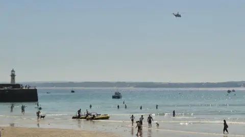 BBC Weather Watcher/Raven A beach at St Ives on a clear sunny day. There are people standing in the sea, swimming and walking on the beach. The coastguard helicopter is flying over the beach. There are a couple of boats moored up in the shallower water and two rigid inflatable boats dragged up onto the pale golden sand. ON the left is the harbour wall which has a white lighthouse on it.