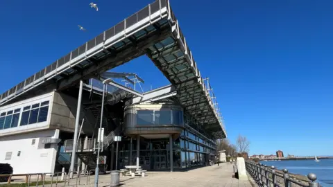 The National Glass Centre is a tall building made of metal panels and glass, overlooking the river Wear. There is seating space and a cafe in front of it, with a dog walker in the distance. Two seagulls are flying above the building.

