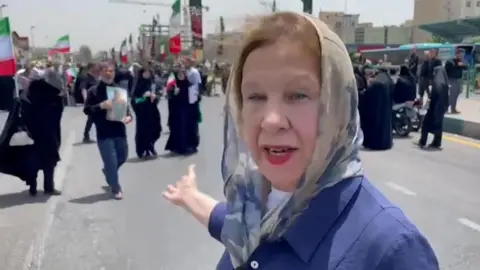 Lyse Doucet in a headscarf and blue shirt pointing at mourners in Iran