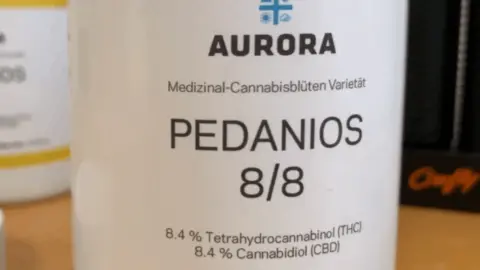 Close-up of a white medical cannabis container labelled “AURORA PEDANIOS 8/8”. The label states “Medizinal-Cannabisblüten Varietät” and lists 8.4% tetrahydrocannabinol (THC) and 8.4% cannabidiol (CBD). The container sits on a wooden surface, with other blurred prescription containers visible in the background.