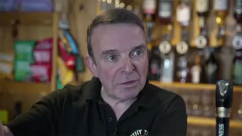 Crown and Anchor landlord David Whitaker wearing a black polo shorts stand behind the bar of the pub