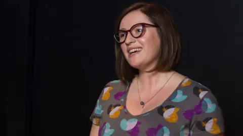 Dr Natalie Diddams A woman with short brown hair and glasses gestures with both hands while speaking on a dark stage. She is wearing a grey patterned dress and a pendant necklace, and is smiling as she looks to one side.
