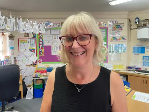Gemma Dawson/BBC School administrator Tracy Wickens. She has a blonde bob with a fringe and is wearing glasses. She is standing in a classroom smiling at the camera.