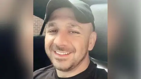 Hemawand Ali Hussein smiling into the camera. He is sat in a car with black seats and is wearing a black cap and t-shirt. 