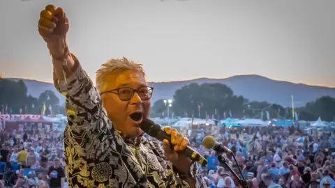 A man with grey hair is facing the camera and is shouting into a microphone on a stage, with his one arm raised in the air. Behind him is a huge crowd of people, and beyond them are lit-up tents, trees and rolling hills