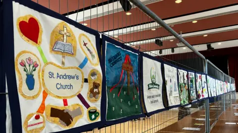 Lots of squares make up a long banner which is attached to metal fencing inside a hall. Each square depicts a different organisation.