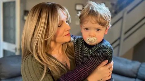 Mother Abbie McNeill has long, blonde hair and is wearing a dark green, V-neck jumper and a silver necklace. She is looking at her son Dalton, who is cuddled into her and has short, wavy, blond hair. Dalton is wearing a purple and green knitted jumper and has a dummy in his mouth.