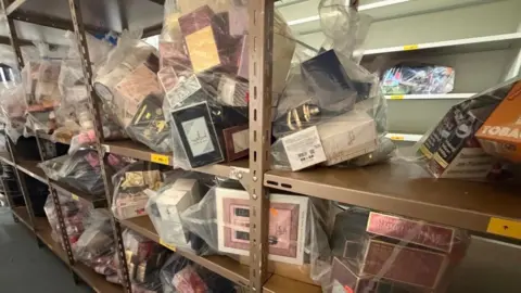 Jasmine Lowe/BBC News Thousands of seized items on shelves at a trading standards warehouse. They are in plastic packaging and are stacked to the ceiling 