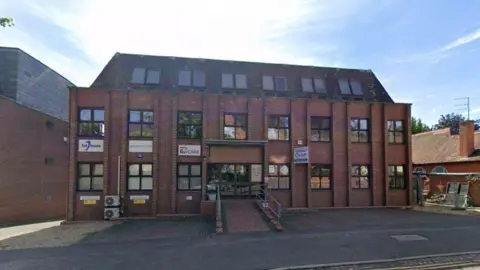 A large three storey red brick building with three storeys of glass windows. 