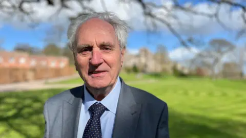 John Hart, former leader of Devon County Council, wearing a light blue shirt, blue patterned tie and grey suit against a blurred background of lawns at Powderham Castle