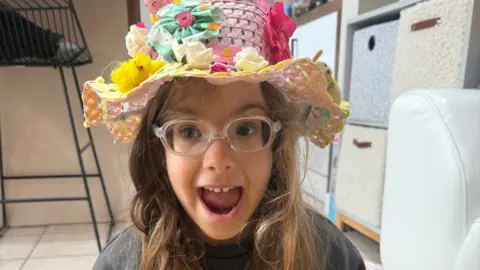A young girl, with her mouth open, looking excited, wearing clear glasses, and an Easter hat on her head, with chicks, flowers and bright colours on her head. She is by boxes in a room with a chair to the left. 