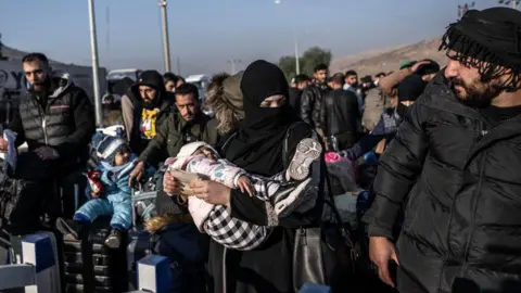 Getty Images Syrian refugees who lives in Turkey wait in a queue at Cilvegozu crossborder gate before entering Syria on 9 December. There are a crowd of men and women holding suitcases. One woman is holding a sleeping child in her arms.