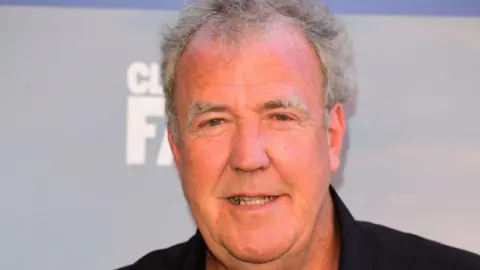 Jeremy Clarkson, a man with short grey hair wearing a black collared shirt, is pictured looking at the camera. Behind him, on a board, are the words "Clarkson's Farm" and "Prime Video". He appears to be at a promotional event.