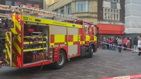 A yellow and red fire engine parked outside a shopping centre and behind a cordon of red and white tape which says 'do not cross'. Bystanders are behind the cordon watching on.