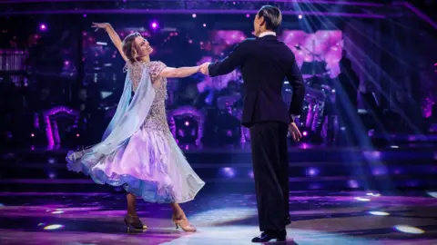BBC Karen Carney, wearing a blue, purple and silver dress, poses mid-dance with her left arm outstretched, while holding the hand of Carlos Gu, who is standing upright while wearing a black suit. They are both on the Strictly Come Dancing dancefloor which is bathed in purple light. A band can be seen performing on their instruments in the background.