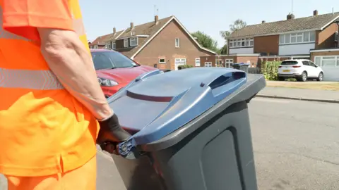 Ant Saddington/BBC A binman wearing an orange high viz jacket and trousers holds onto a rubbish bin with a blue lid.