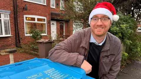 Councillor Charles Quinn next to a blue bin. He is wearing a Santa hat and smiling at the camera. He has ginger facial hair and a small stubble.