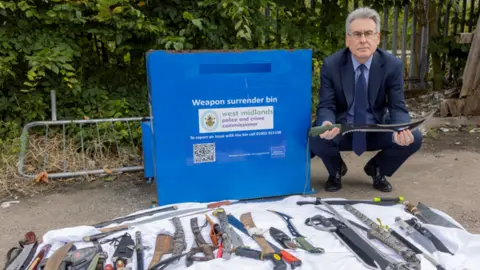 Simon Foster, a man with silver hair wearing a blue suit, squats next to a blue weapons disposal bin. In front of the bin is white cloth laid out on the ground with dozens of knives and weapons on it.