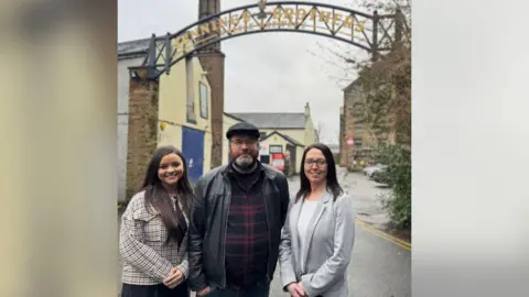 Chris France (middle) with flat cap and beard and leather jacket and Rebecca Canfield (right) with grey cardigan and glasses and dark brown hair. There is a woman to the left of the pair wearing a chequered jacket. 