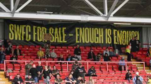 PA Media Black banner that says 'SWFC for sale - enough is enough' at the back of a stadium with red seats where numerous people sit