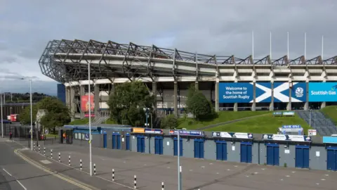 An exterior view of Murrayfield Stadium in Edinburgh. There is a row of turnstiles with blue doors that are closed. Behind the turnstiles is a sloped grassy area and then the stadium with its exposed steel work on its roof. There is a sign with a Saltire and the message: "Welcome to the home of Scottish Rugby".