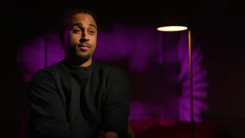 Sebastian Wray sits in a studio interview, wearing a dark sweatshirt, with purple lighting and a floor lamp behind him.
