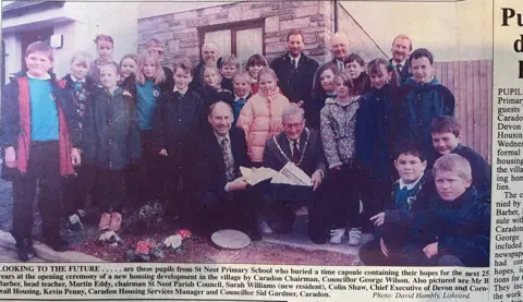 An article in a newspaper showing primary school children with notable members of the parish council and the head teacher with a box and letter going inside.