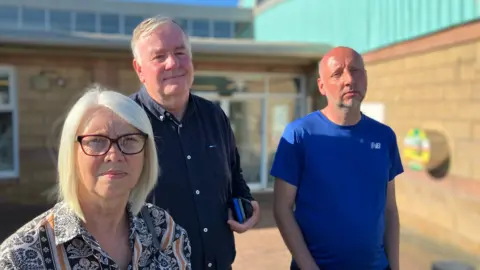 BBC Three participants in the Grief Cafe, one woman and two men, standing outside the main entrance of the Oval Centre in Workington on a sunny afternoon.