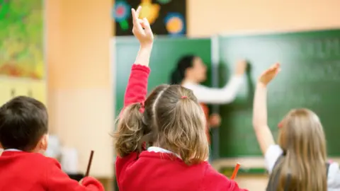 A classroom. A schoolgirl in a red cardigan raises her hand to answer a teacher's question. The teacher writes on a board in the background.