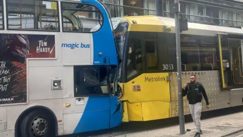 A blue-and-white double-decker Stagecoach bus is pressed against the front of a yellow Metrolink tram with a shattered windscreen. The front of the bus is also crumpled. A policeman talks into his walkie-talkie while walking in front of the crashed vehicles in Piccadilly Gardens in Manchester city centre.