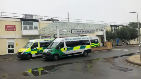 Main entrance sign at Frimley Park Hospital, ambulance and patient transport service vehicles outside.