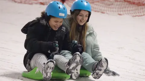 Caring Together Two young girls are sat on green sledges and travelling down a snowy slope. Their feet are off the ground and covered in snow and both are laughing as they go. They wear blue helmets and warm winter coats.