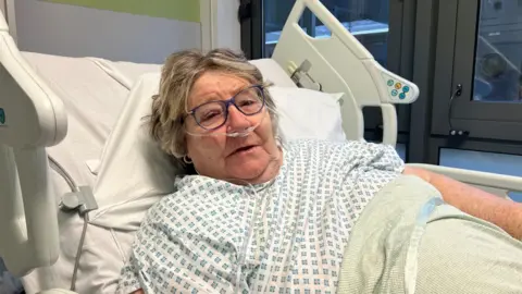 A woman with short grey hair and glasses is lying in a hospital bed in a hospital gown with an oxygen tube in her nose. She is wearing a hospital gown and is lying under covers.