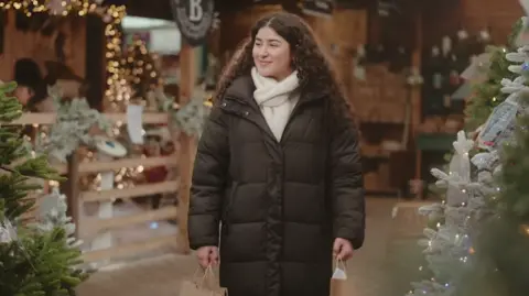 North Northamptonshire Council A woman in a black coat with long dark curly hair holding two paper shopping bags in a Christmas-themed setting with fairy lights and Christmas trees.