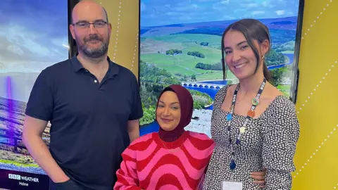 Andrew Evans, wearing a blue polo shirt, stands with Rima Ahmed, who wears a puple headscarf and a pink and red striped jump, and Alice Toon, who wears a spotty dress. The three are all smiling together.