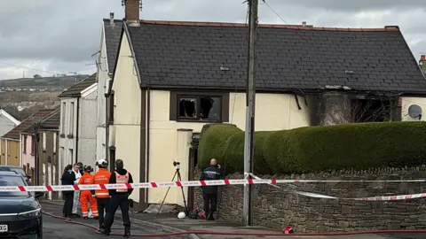 Investigators gather in the street by a house where first-floor windows have been either shattered or are missing and the yellow painted external walls have been partially blackened as if by fire damage