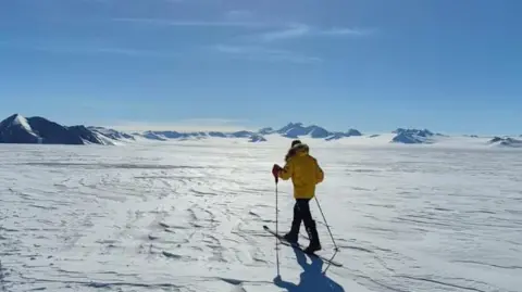 Jonny Huntington A man with skis trekking into the snow on a clear day.