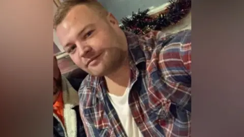 Selfie of Ben Crook. He wears a white t-shirt and a red and navy flannel overshirt. He smiles at the camera and has short blonde hair. 