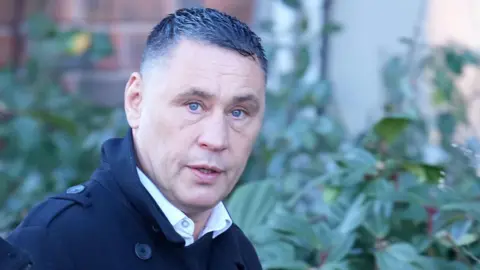 A man with short black hair wearing a black jacket. He is walking past a hedge and looking into the camera to his right. He look surprised.