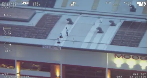 Staffordshire Police Surveillance footage marked with white date stamps and location data shows the roof of a building with a home-made Free Palestine banner hanging off the side, some way left of the site's Moog branding. On the roof itself, three people are visible sitting and standing on a white roof panel near ventialtion shafts.